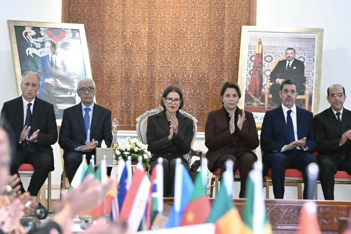 SAR la Princesse Lalla Meryem assise lors de la cérémonie de clôture du Forum Africain du Parlement de l’Enfant, applaudissant, encadrée par les portraits de feu Hassan II et de SM le Roi Mohammed VI, avec en arrière-plan de petits drapeaux africains.